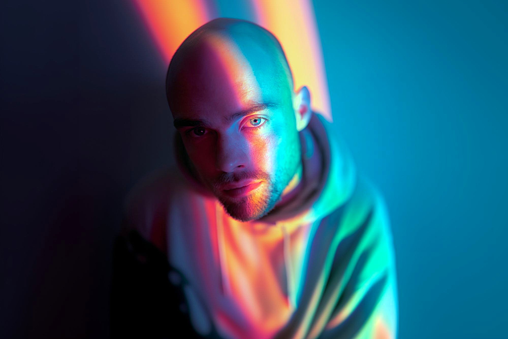 A man in a hoodie with a shaved head is illuminated by vibrant blue, pink, and orange lights, casting colorful shadows on his face and background in a dramatic, artistic portrait.