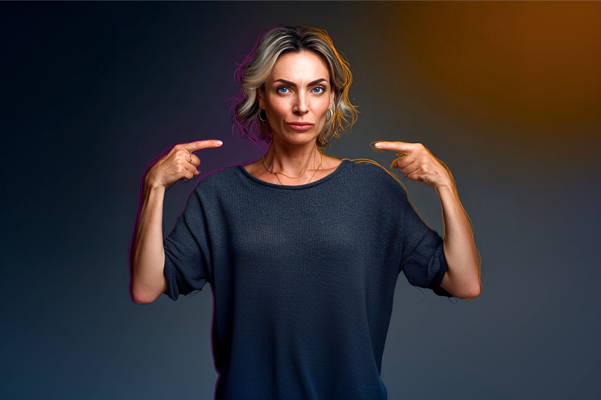 A woman with short blonde hair and a serious expression stands against a dark background, pointing at herself with both hands. She wears a loose dark gray top.