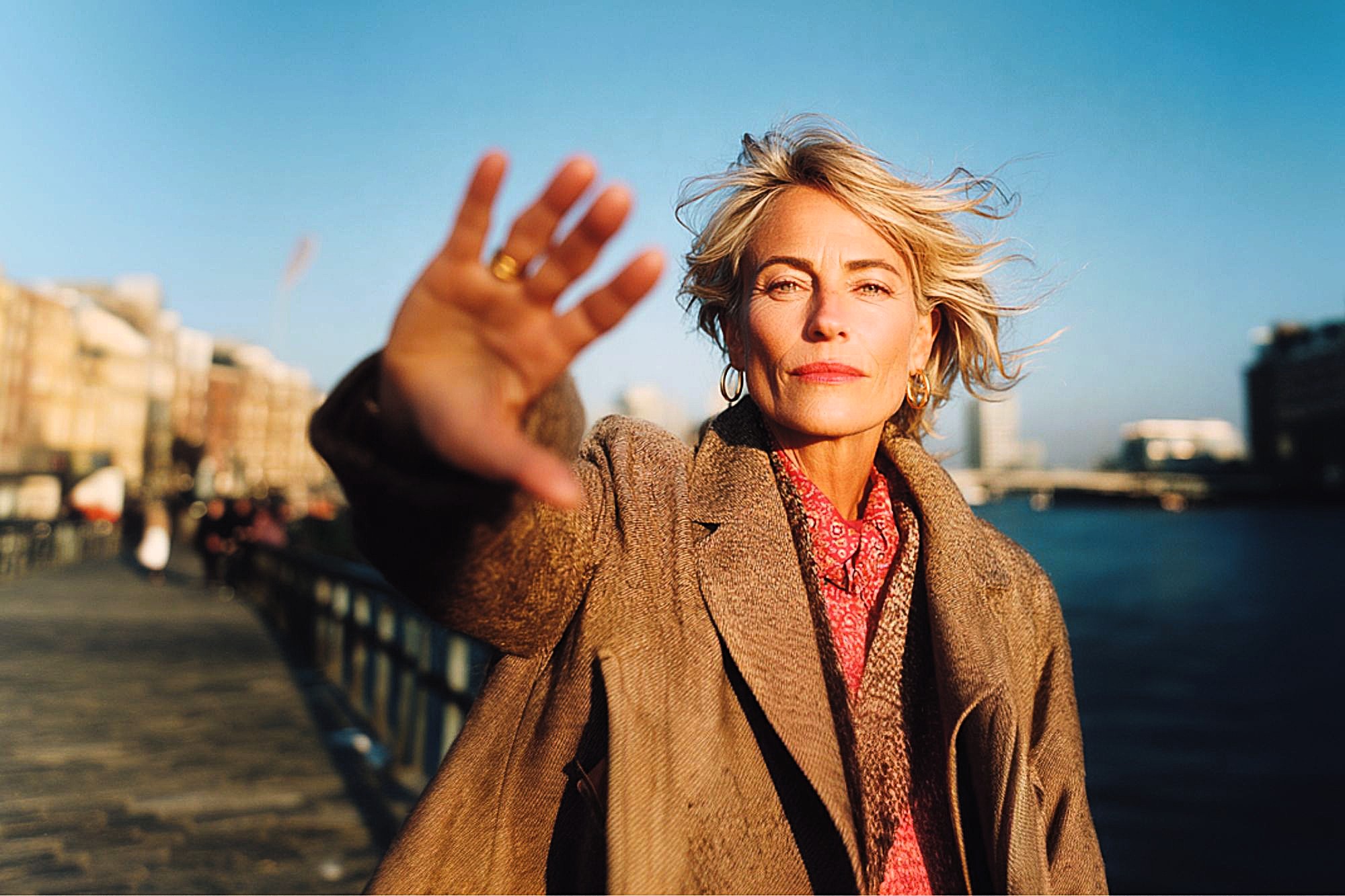 A woman with blonde hair and a brown coat stands on a riverside walkway, facing the camera with her hand outstretched, partially covering her face. The background shows water, buildings, and a clear blue sky.