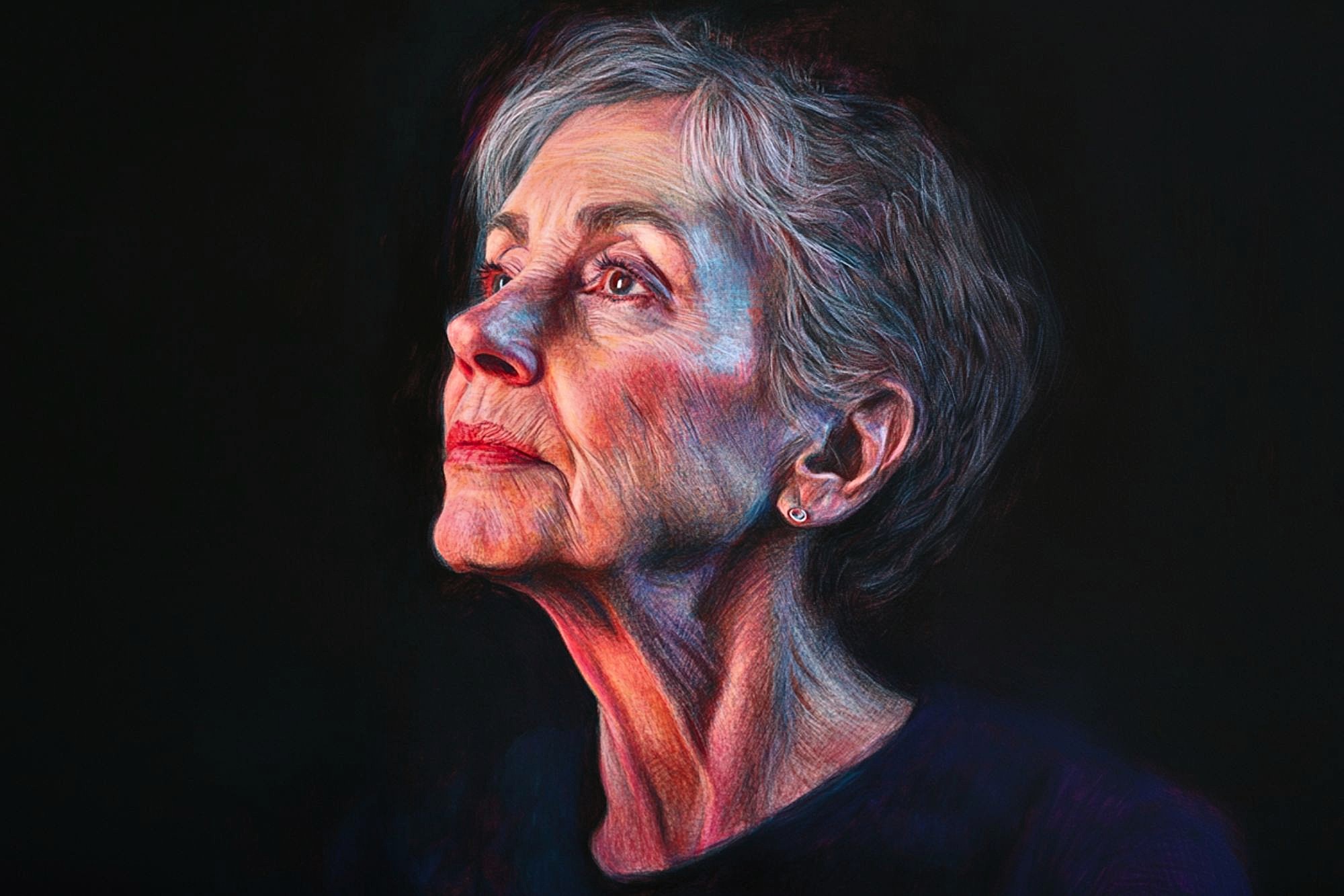 A mature woman with short gray hair gazes upward in soft, colorful lighting against a dark background, her expression thoughtful and serene. She wears a simple, dark top and small stud earrings.