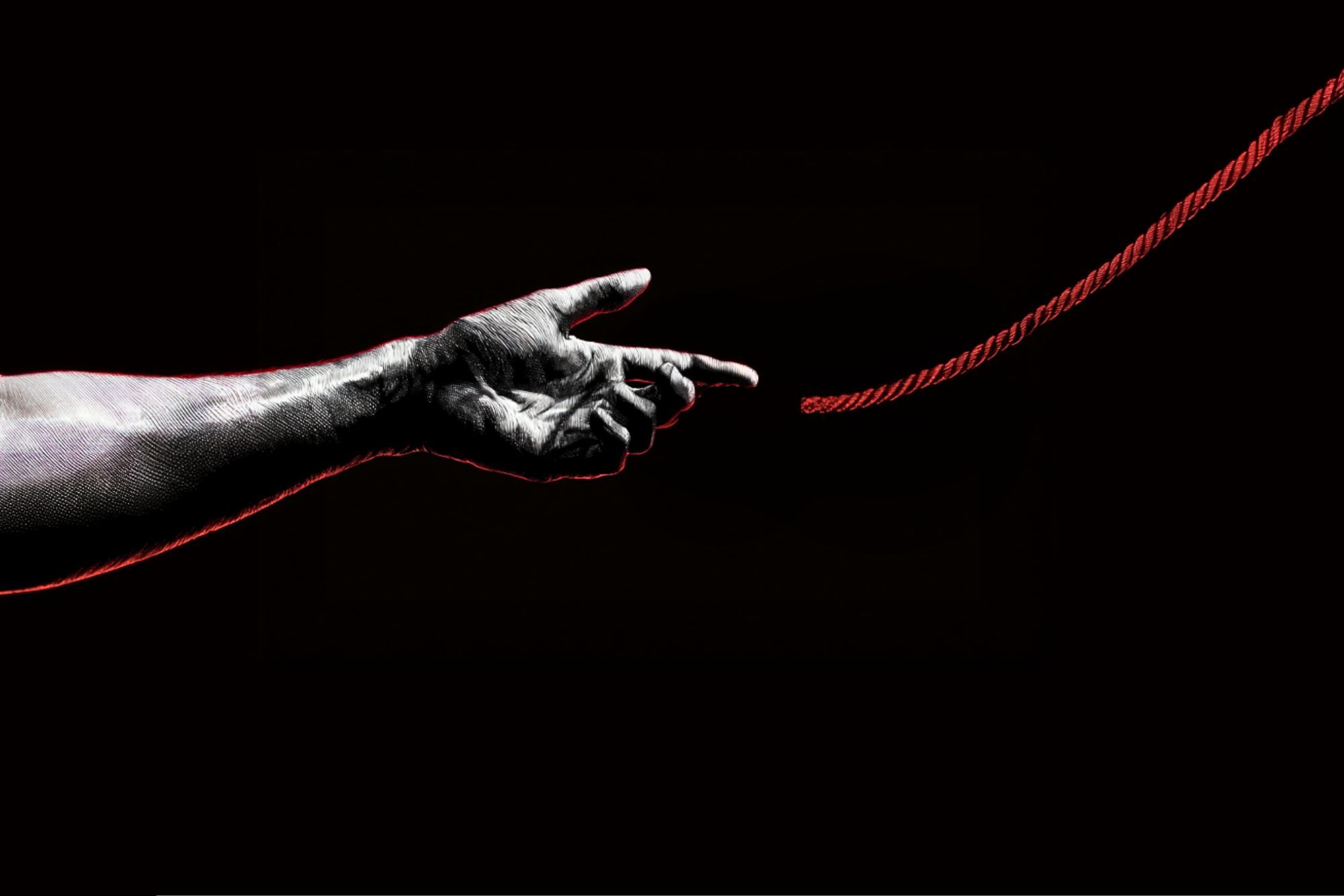 A grayscale hand stretches toward a red rope against a black background, with the fingers almost touching the rope, symbolizing connection or reaching out.