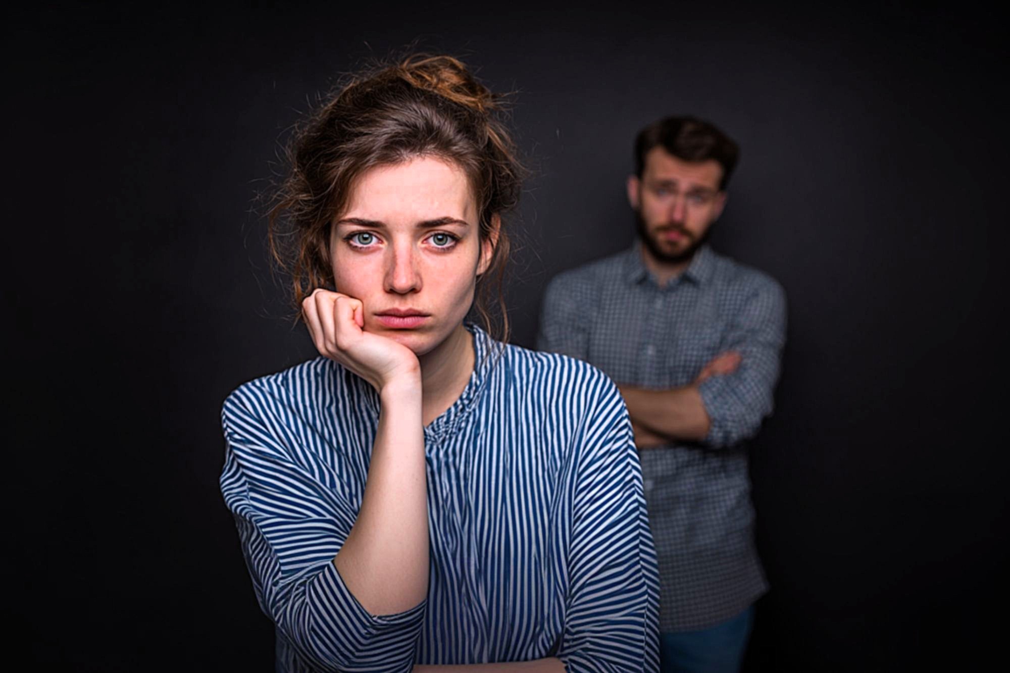 A woman with a sad expression rests her chin on her hand in the foreground, while a man with crossed arms stands in the blurred background against a dark backdrop.