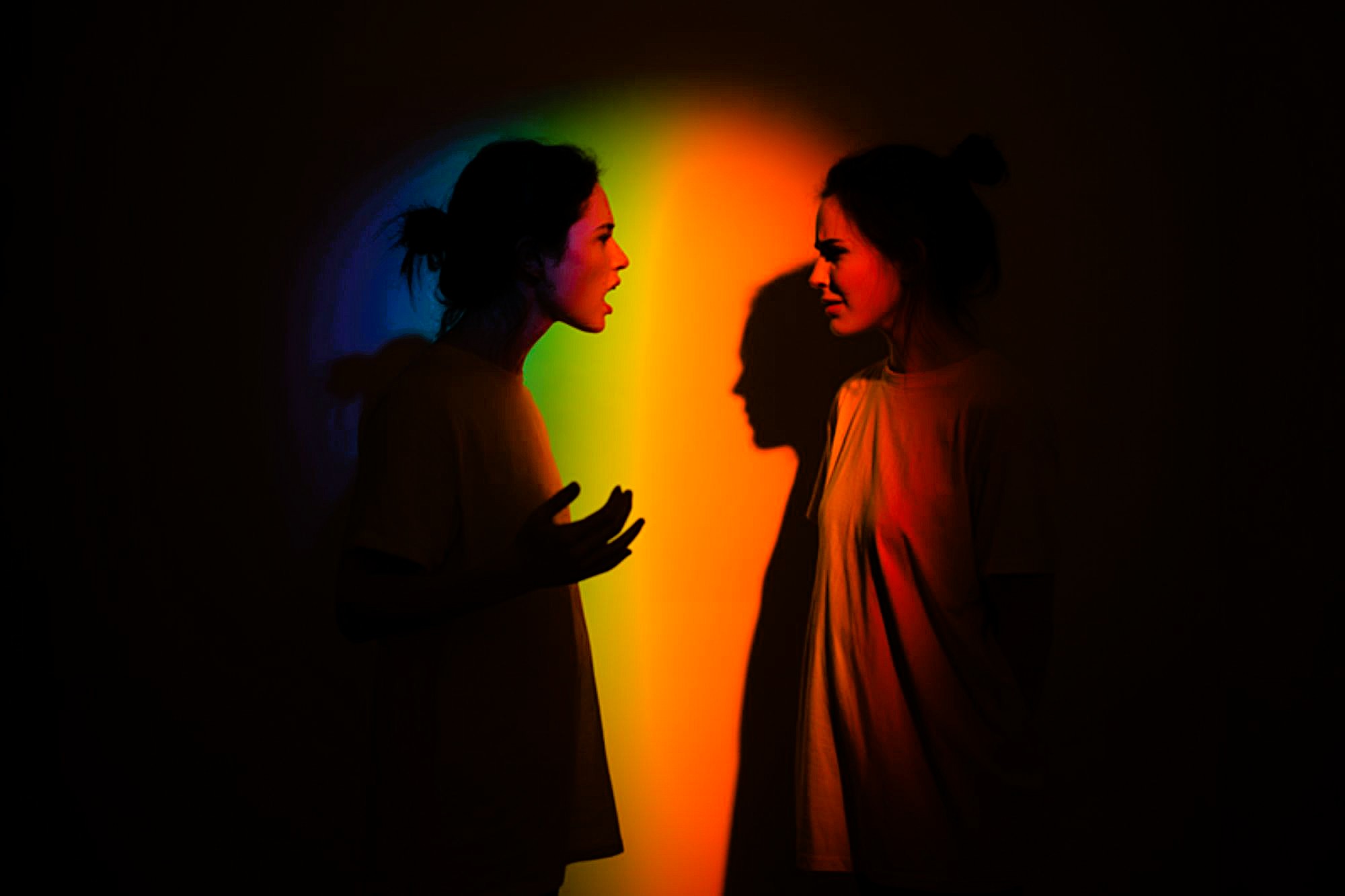 Two women face each other in a dark room, standing in front of a colorful rainbow light. One woman gestures and appears to be yelling, while the other looks upset. Both cast shadows on the wall behind them.
