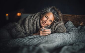 A woman with gray hair, wrapped in a thick knit blanket, smiles warmly while holding a mug. She is lying on a bed with cozy gray bedding, with candles softly glowing in the background.