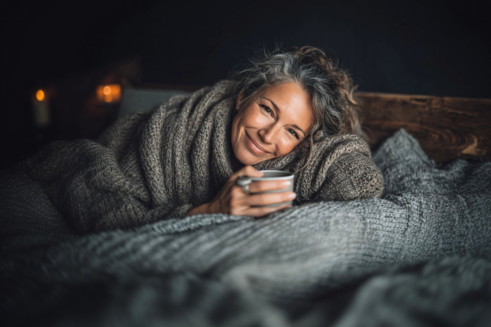 A woman with gray hair, wrapped in a thick knit blanket, smiles warmly while holding a mug. She is lying on a bed with cozy gray bedding, with candles softly glowing in the background.