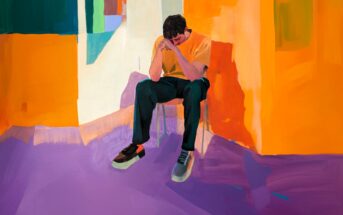 A person sits on a chair in a brightly colored room with orange and purple tones, head bowed and hands covering their face, suggesting a mood of sadness or contemplation.