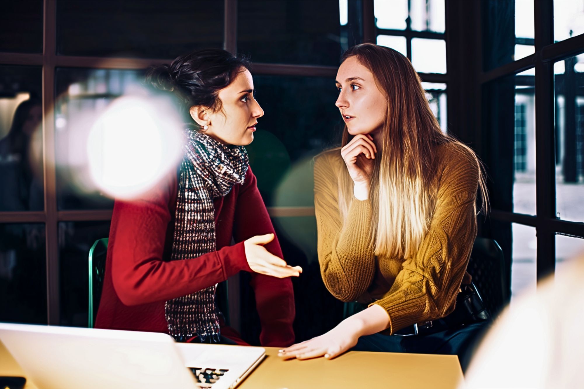 Two women sit at a table in conversation, one gesturing while speaking, the other listening with her hand on her chin. They are indoors, with a laptop on the table and light streaming in through windows.