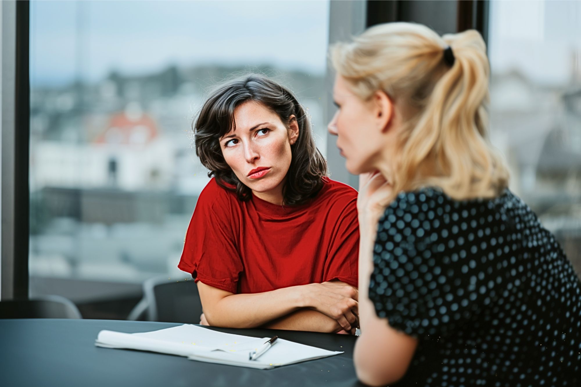Two women sit at a table with papers and a pen. One woman, wearing a red shirt, looks skeptically at the other, who is in a black polka dot top. They appear to be having a serious conversation indoors.