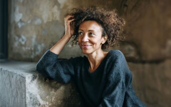 A woman with curly hair wearing a dark sweater sits by a concrete ledge, smiling and looking upward. The background is a textured, rustic wall. The lighting is natural and soft.