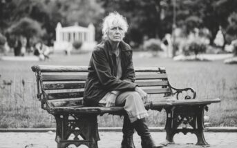 An older woman with white hair sits alone on a park bench, wearing a dark jacket and boots. The scene is in black and white, with trees and a gazebo blurred in the background.