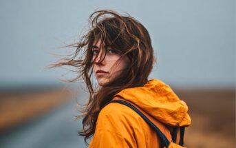 A person with long brown hair blown by the wind wears a yellow rain jacket and backpack, standing on a deserted road with a blurred, overcast landscape in the background.