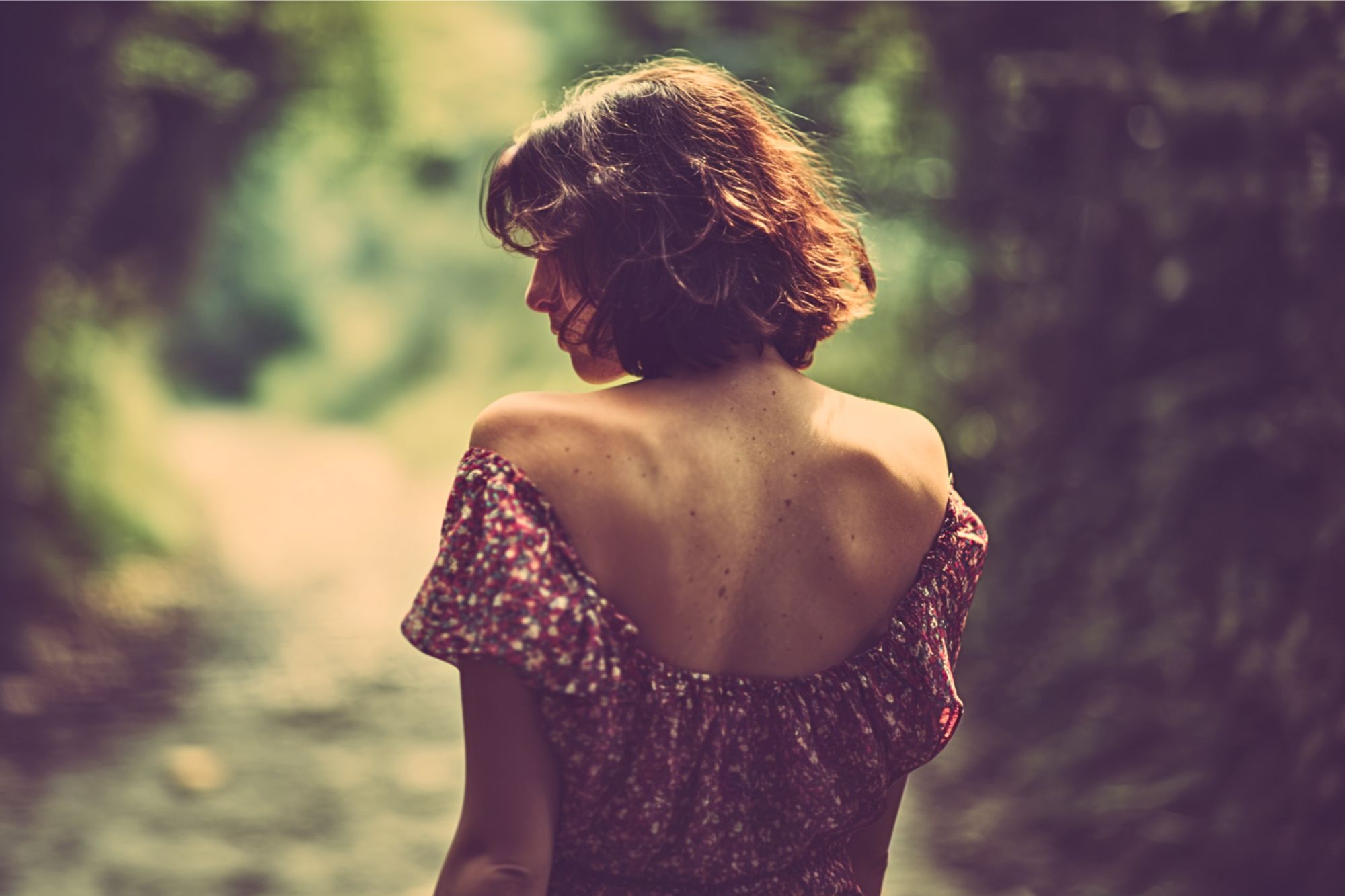 A woman with short brown hair in a floral off-shoulder dress stands on a sunlit path, facing away and looking over her shoulder. The background is blurred with greenery, creating a soft, dreamy atmosphere.