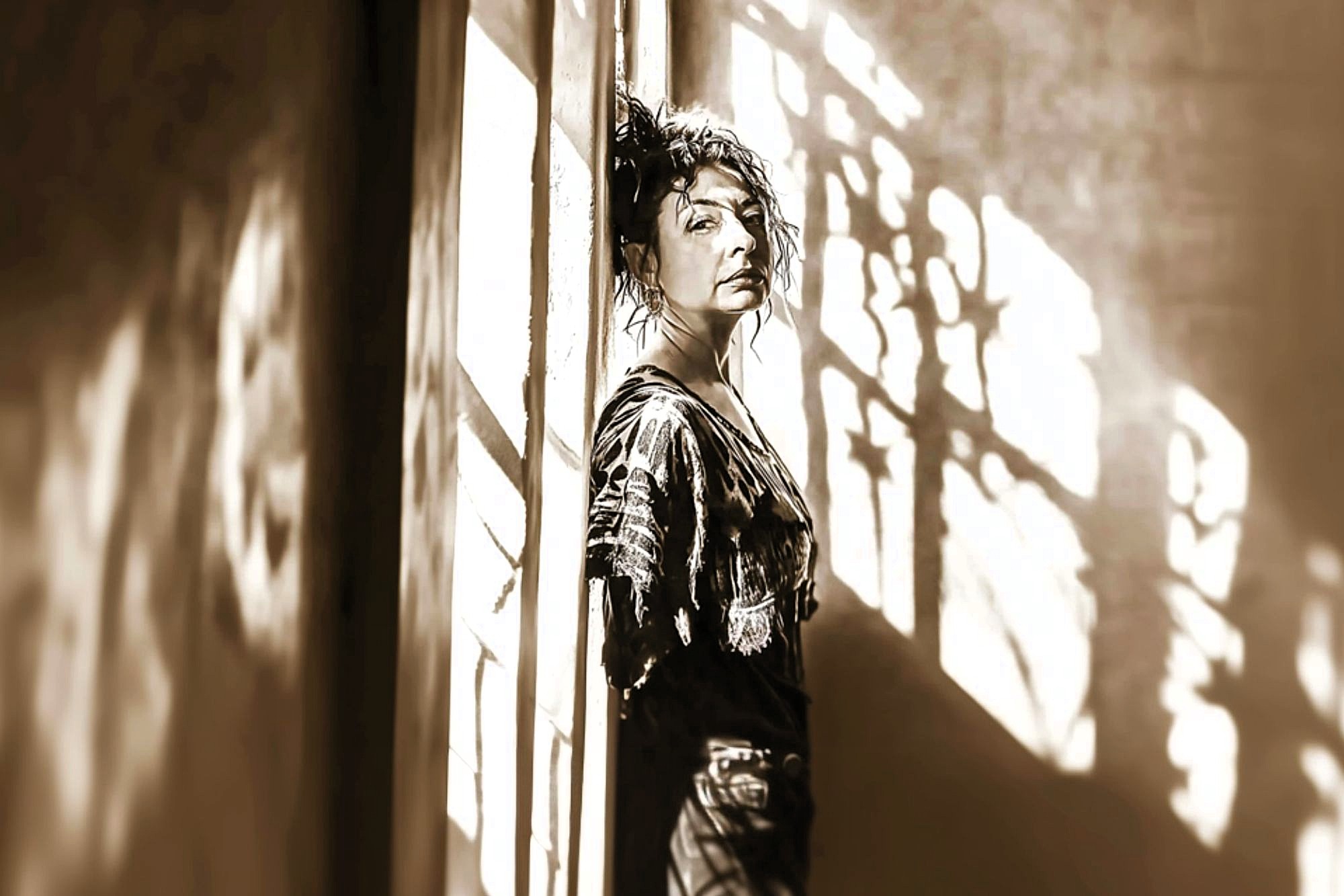 A person with curly hair leans against a wall in a narrow hallway, illuminated by dramatic sunlight and shadows streaming through a patterned window. The scene is in sepia tones, giving it a vintage feel.