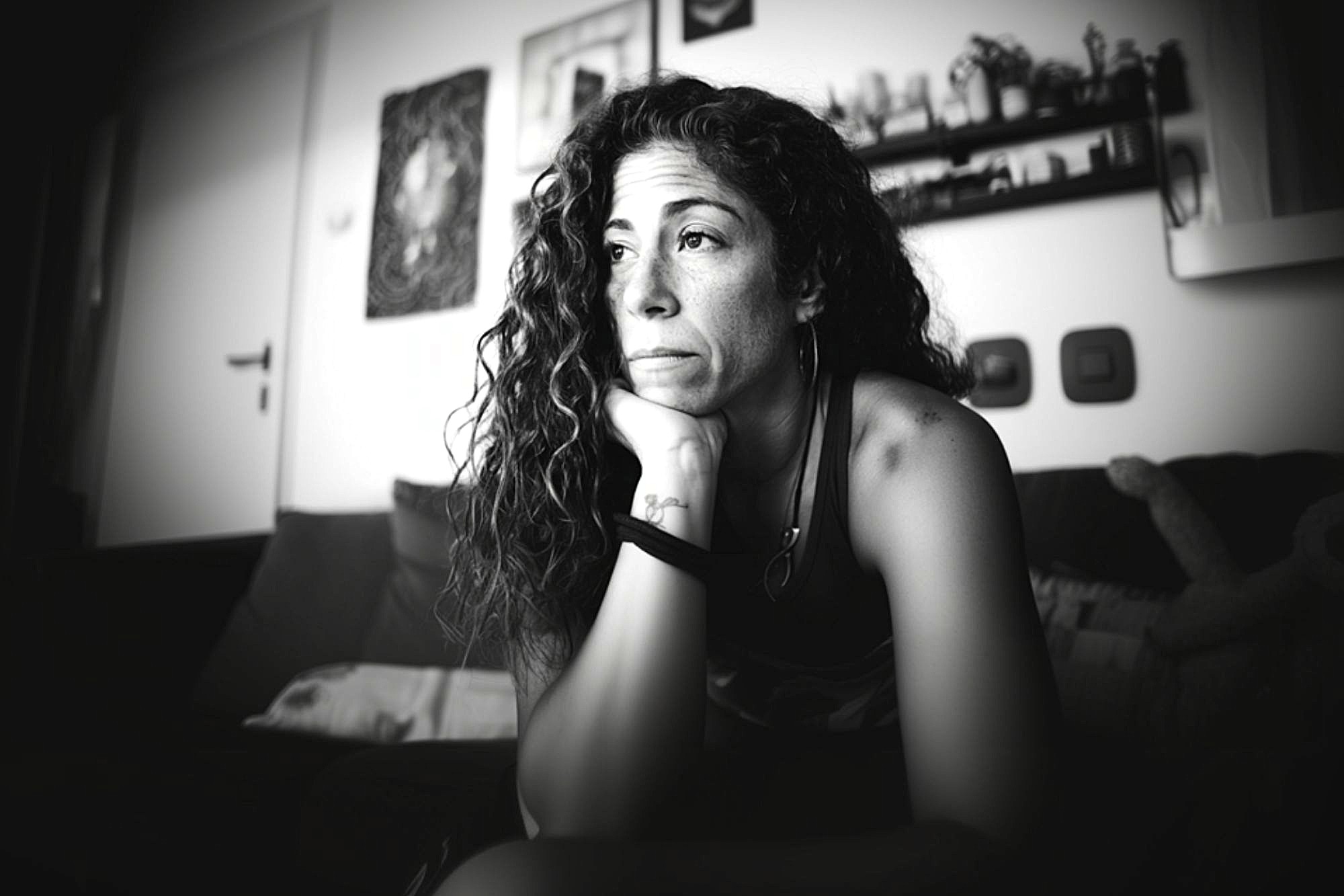 A woman with long curly hair sits on a couch, resting her chin on her hand and gazing thoughtfully into the distance in a softly lit, monochrome room with shelves and framed art on the wall behind her.