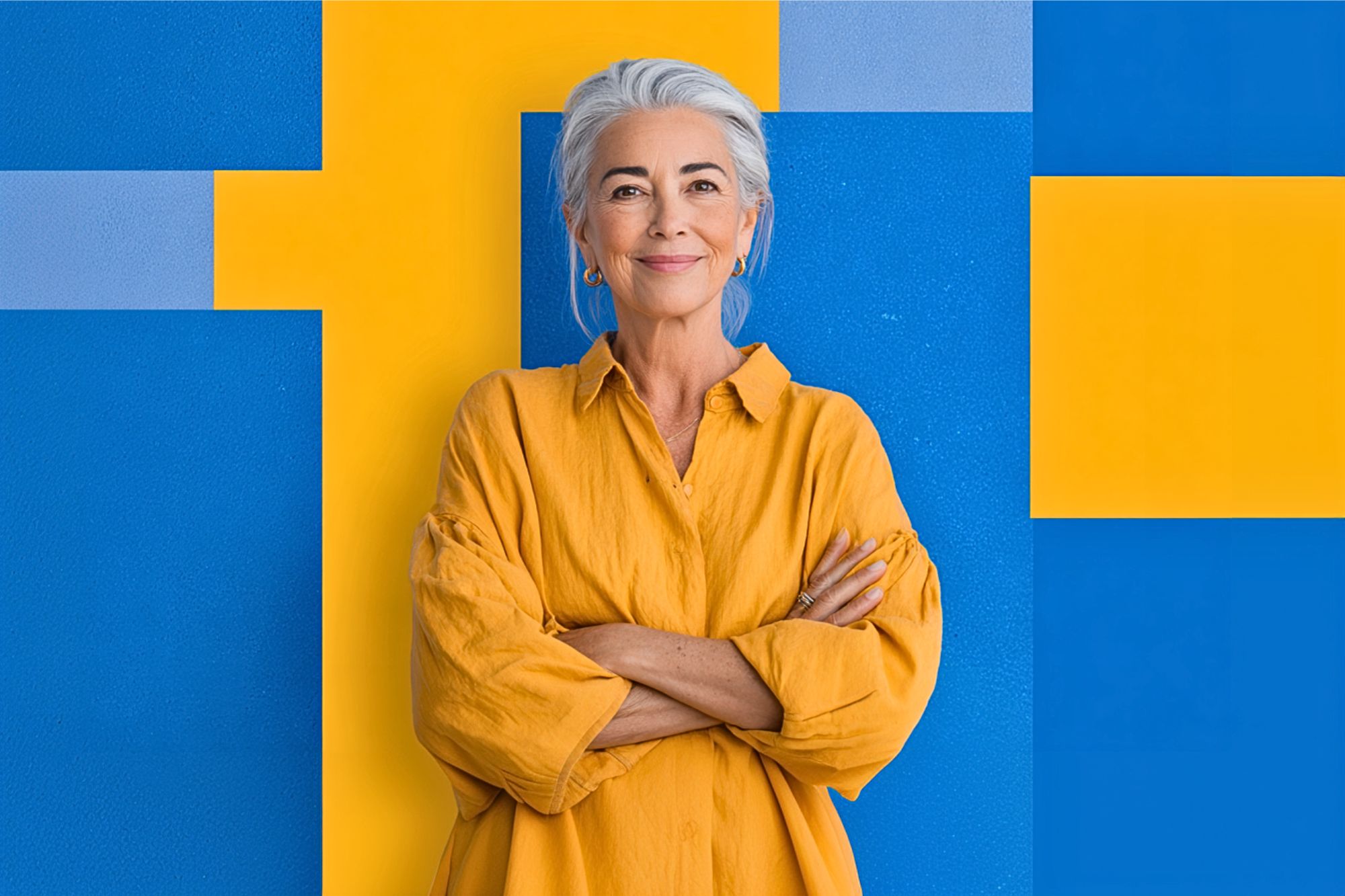 A smiling older woman with gray hair and a yellow shirt stands with arms crossed in front of a bold geometric background of yellow and blue rectangles.