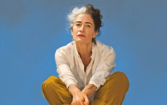 A woman with short, curly gray and dark hair sits cross-legged, wearing a white shirt and mustard yellow pants, against a clear blue background, looking thoughtfully at the camera.