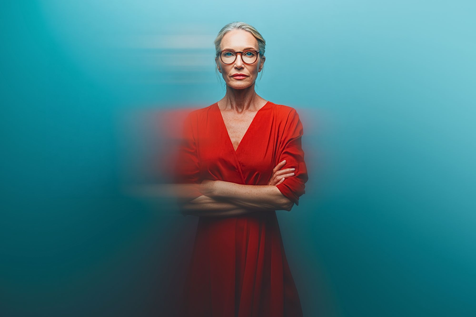 A confident older woman with gray hair in a bun and red glasses stands with arms crossed, wearing a red dress against a smooth blue background with a motion blur effect around her.