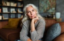 An older woman with long gray hair sits on a brown sofa, resting her chin on her hand and looking thoughtfully at the camera. A bookshelf and cushions are visible in the background, giving a cozy, homey feel.