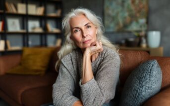 An older woman with long gray hair sits on a brown sofa, resting her chin on her hand and looking thoughtfully at the camera. A bookshelf and cushions are visible in the background, giving a cozy, homey feel.