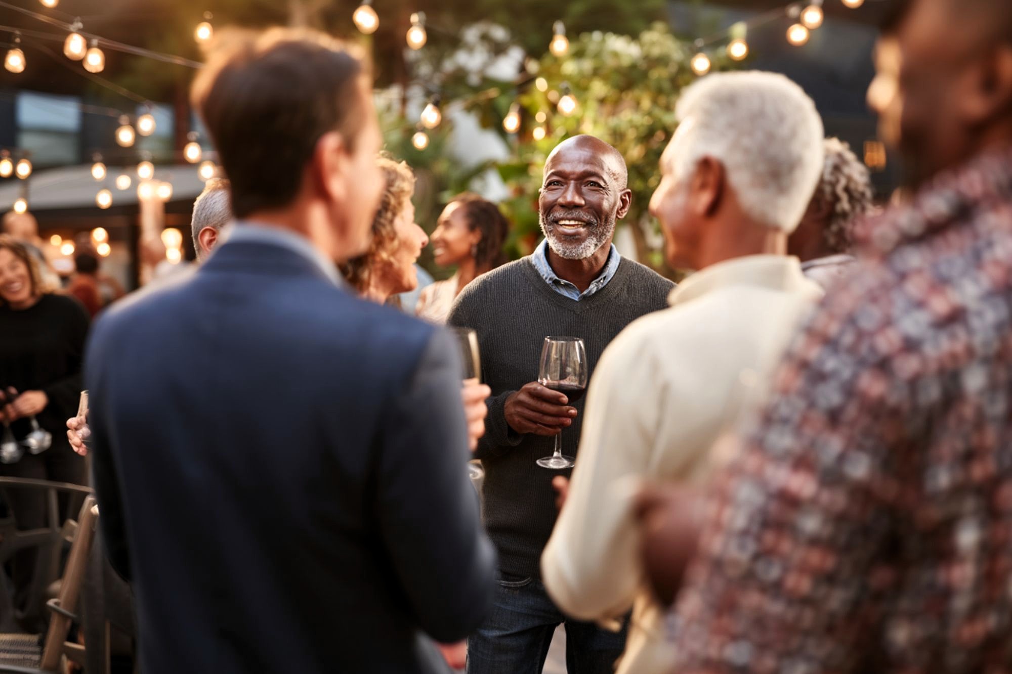 A group of people socializing outdoors at a party or gathering, with string lights overhead. A man in the center smiles while holding a wine glass, engaging in conversation with others.
