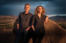 A man and woman stand outdoors near a large rock, with mountains and an open landscape in the background. Both wear dark shirts and have relaxed, confident expressions in the warm, late-day sunlight.