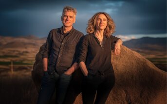 A man and woman stand outdoors near a large rock, with mountains and an open landscape in the background. Both wear dark shirts and have relaxed, confident expressions in the warm, late-day sunlight.