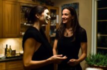 Two women stand in a warmly lit kitchen; one is smiling while the other wears a white theatrical mask and holds a drink. Wine bottles and herbs are visible in the background.