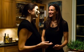 Two women stand in a warmly lit kitchen; one is smiling while the other wears a white theatrical mask and holds a drink. Wine bottles and herbs are visible in the background.