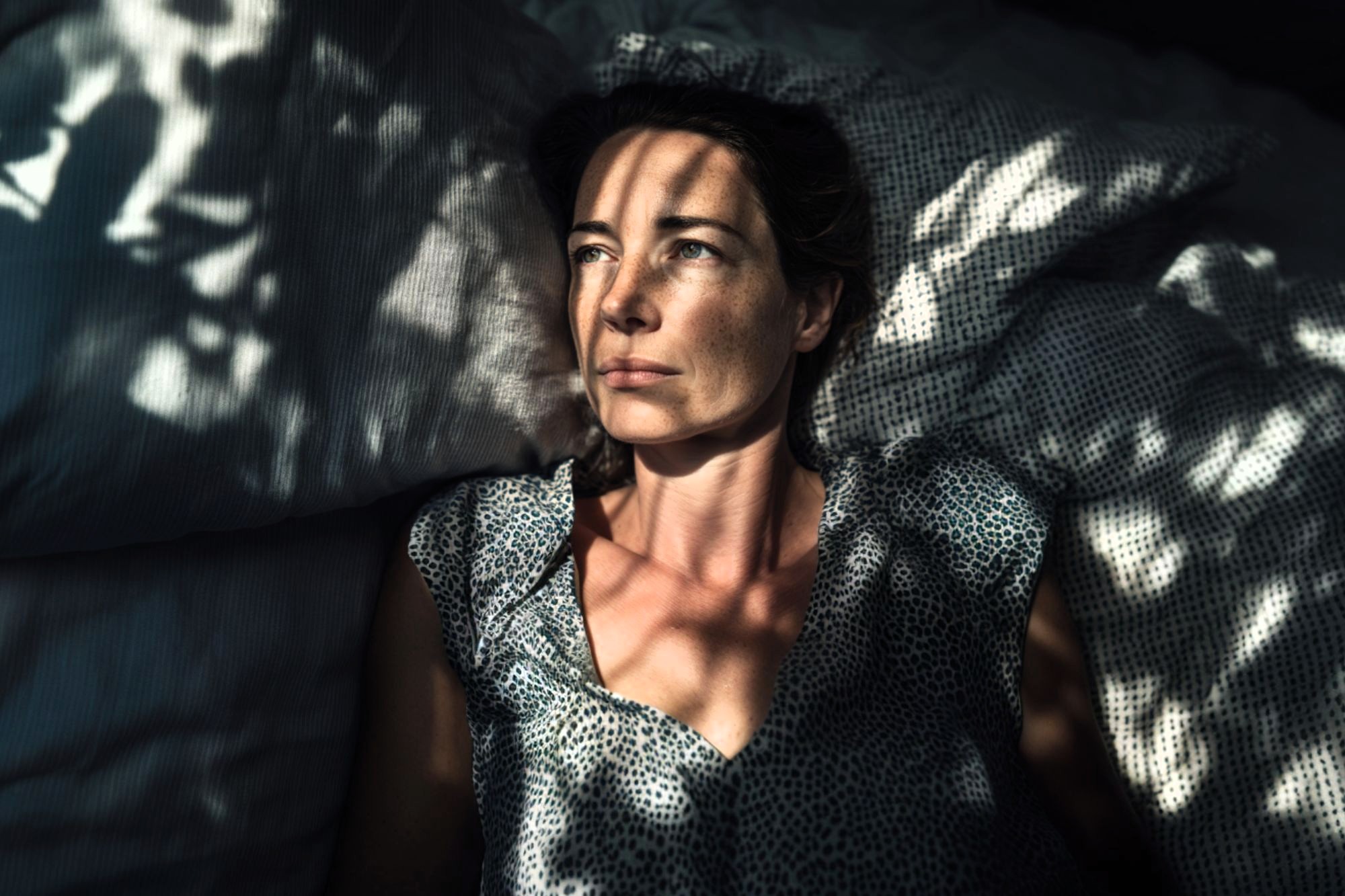 A woman lies on a bed, looking upward with a thoughtful expression. Dappled sunlight and shadows from leaves create patterns across her face, pillow, and shirt.