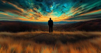 A person stands in tall golden grass on a hilltop, gazing at a vibrant, colorful sunset sky filled with swirling clouds and stars over a vast, mountainous landscape.