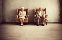 An elderly woman and man sit separately in vintage armchairs against a worn, gray wall, both dressed formally and looking straight ahead. The setting appears quiet and sparse, with empty space around them.