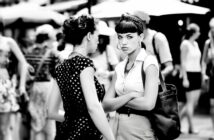 Two women stand close together in a busy outdoor setting. One faces the camera with arms crossed and a serious expression, while the other, seen from the side, appears to be talking. The background is blurred with people and umbrellas.