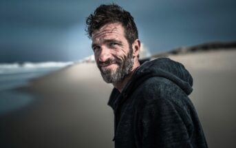 A middle-aged man with a beard and tousled hair, wearing a dark hoodie, stands on a sandy beach under a cloudy sky, smiling slightly at the camera. The ocean and dunes are blurred in the background.