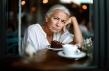An older woman with gray hair, wearing a white blouse, sits at a table in a café, resting her head on her hand and looking thoughtful. A slice of chocolate cake and a cup of coffee are in front of her.