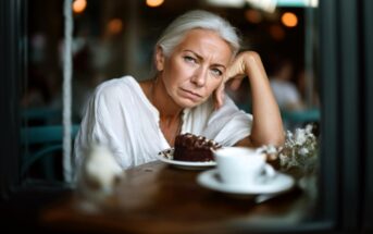 An older woman with gray hair, wearing a white blouse, sits at a table in a café, resting her head on her hand and looking thoughtful. A slice of chocolate cake and a cup of coffee are in front of her.