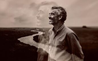 A smiling man stands in a field near a winding river, with a transparent, larger image of himself in the background, creating a dreamy, reflective atmosphere. The image is in sepia tones.
