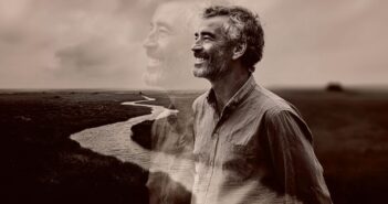 A smiling man stands in a field near a winding river, with a transparent, larger image of himself in the background, creating a dreamy, reflective atmosphere. The image is in sepia tones.