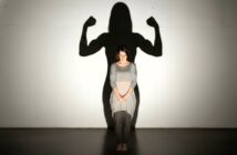 A woman stands barefoot with her head bowed, casting a shadow on the wall behind her that shows her in a strong, flexed-arm pose, symbolizing inner strength and confidence.