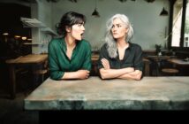 Two women sit at a table in a rustic café. The younger woman, with dark hair in a green blouse, looks surprised with her mouth open. The older woman with gray hair and folded arms looks serious and unimpressed.